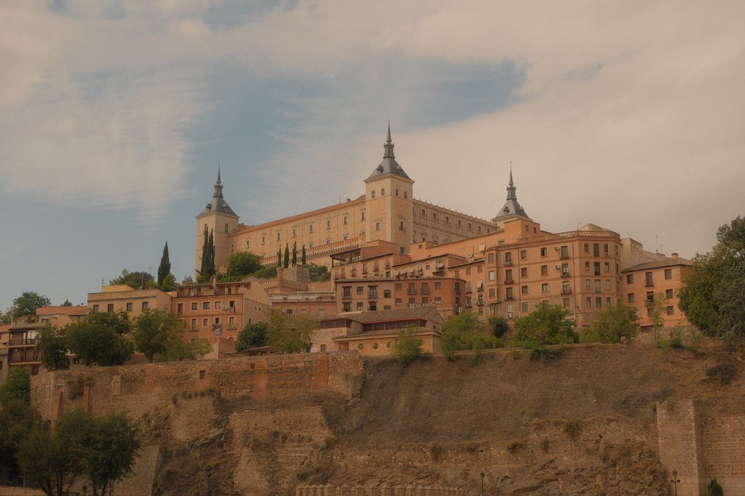 A sandstone palace with slate-roofed towers on a hill above old town houses, under a soft cloudy sky