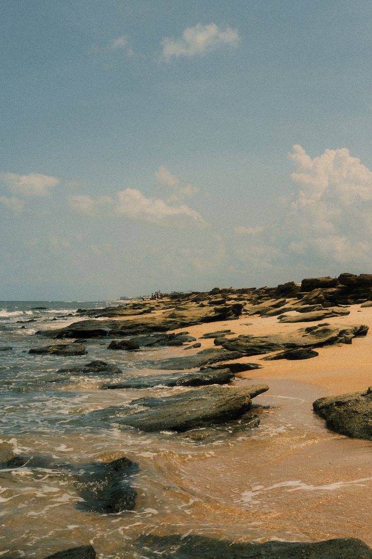 Dark rocks and a wave washing over a sandy beach under a pale blue sky