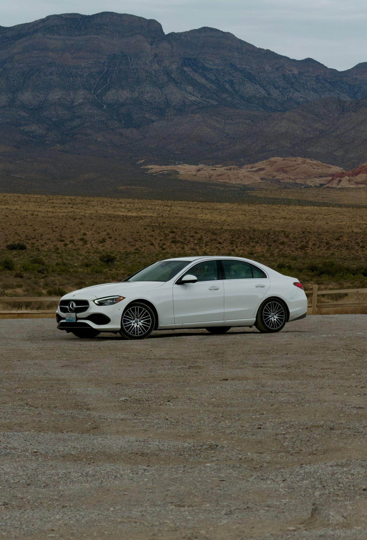 A white sedan parked on gravel in front of a desert plain and dark mountains