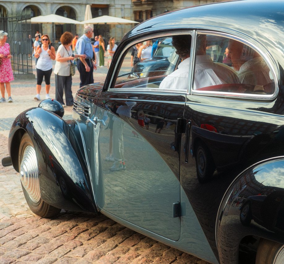 A glossy black vintage car on cobblestones, reflecting the plaza and onlookers